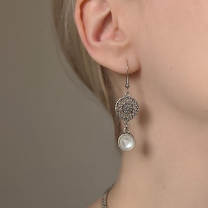 May include: A silver-toned drop earring featuring a circular filigree charm with an intricate openwork pattern, suspended above a small, round, white cabochon stone. The earring hangs from a simple silver-toned hook wire.