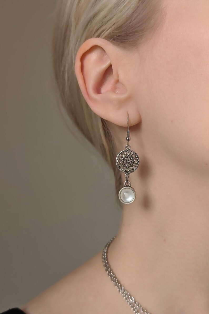 May include: A silver-toned drop earring featuring a circular filigree charm with an intricate openwork pattern, suspended above a small, round, white cabochon stone. The earring hangs from a simple silver-toned hook wire.