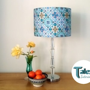 May include: A table lamp with a patterned lampshade in blue, teal, and white geometric design. The lamp has a clear, stacked base and a silver-coloured metal base. A small vase with yellow roses and a bowl of oranges are on the table.