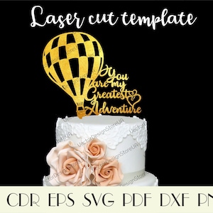 May include: Gold glitter cake topper with a hot air balloon and the text "You are my greatest adventure". The topper is on a white cake with peach roses.