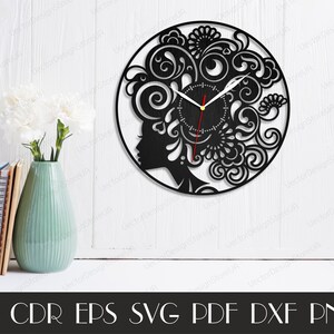 Puede incluir: Reloj en blanco y negro con una silueta de rostro de mujer y dise&ntilde;os florales intrincados. La esfera del reloj es redonda y tiene un fondo negro con manecillas blancas.