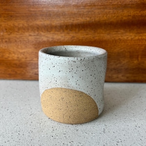 May include: A speckled white ceramic tumbler with a tan, rounded accent on the lower half. The handmade cup has a cylindrical shape and is suitable for beverages. The cup is displayed on a light-colored surface.
