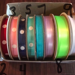 K&ouml;nnte beinhalten: Eine Schachtel mit acht Rollen Band. Die B&auml;nder sind in verschiedenen Farben und Mustern, darunter wei&szlig;e Punkte auf einem lila Band, wei&szlig;e Punkte auf einem roten Band, wei&szlig;e Punkte auf einem t&uuml;rkisfarbenen Band und ein einfarbiges hellrosa Band. Die B&auml;nder sind von 1 bis 9 nummeriert.