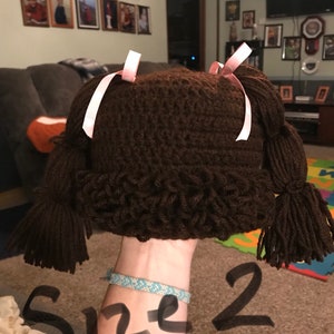 K&ouml;nnte beinhalten: Braune H&auml;kelm&uuml;tze mit zwei braunen Garnz&ouml;pfen und rosa B&auml;ndern. Die M&uuml;tze ist mit "Gr&ouml;&szlig;e 2" gekennzeichnet.