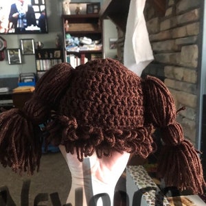 K&ouml;nnte beinhalten: Ein brauner H&auml;kelhut mit zwei Z&ouml;pfen. Der Hut ist so konzipiert, dass er wie eine Per&uuml;cke aussieht.