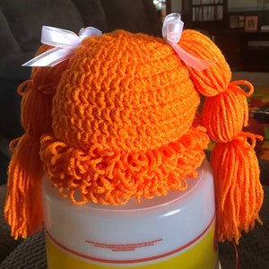 K&ouml;nnte beinhalten: Orangefarbenes geh&auml;keltes Puppenhaar mit zwei wei&szlig;en Schleifen und langen orangen Garnz&ouml;pfen.