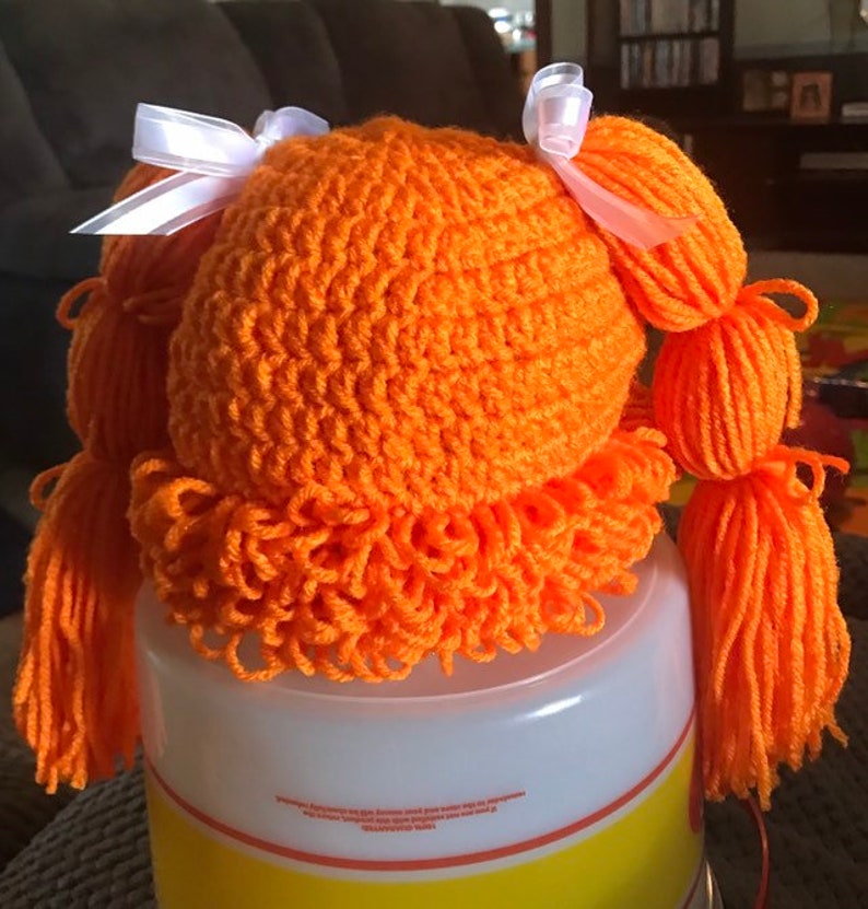 K&ouml;nnte beinhalten: Orangefarbenes geh&auml;keltes Puppenhaar mit zwei wei&szlig;en Schleifen und langen orangen Garnz&ouml;pfen.