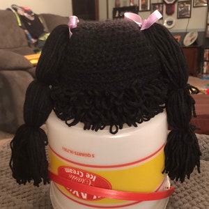 K&ouml;nnte beinhalten: Ein schwarzer Garn-Puppenkopf mit zwei Z&ouml;pfen, die mit lila B&auml;ndern gebunden sind. Der Puppenkopf sitzt auf einem wei&szlig;en Plastik-Eisbeh&auml;lter mit einem gelben Etikett, auf dem "Classic Ice Cream 5 Quarts 1.473L" steht.