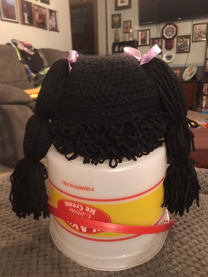 K&ouml;nnte beinhalten: Ein schwarzer Garn-Puppenkopf mit zwei Z&ouml;pfen, die mit lila B&auml;ndern gebunden sind. Der Puppenkopf sitzt auf einem wei&szlig;en Plastik-Eisbeh&auml;lter mit einem gelben Etikett, auf dem "Classic Ice Cream 5 Quarts 1.473L" steht.