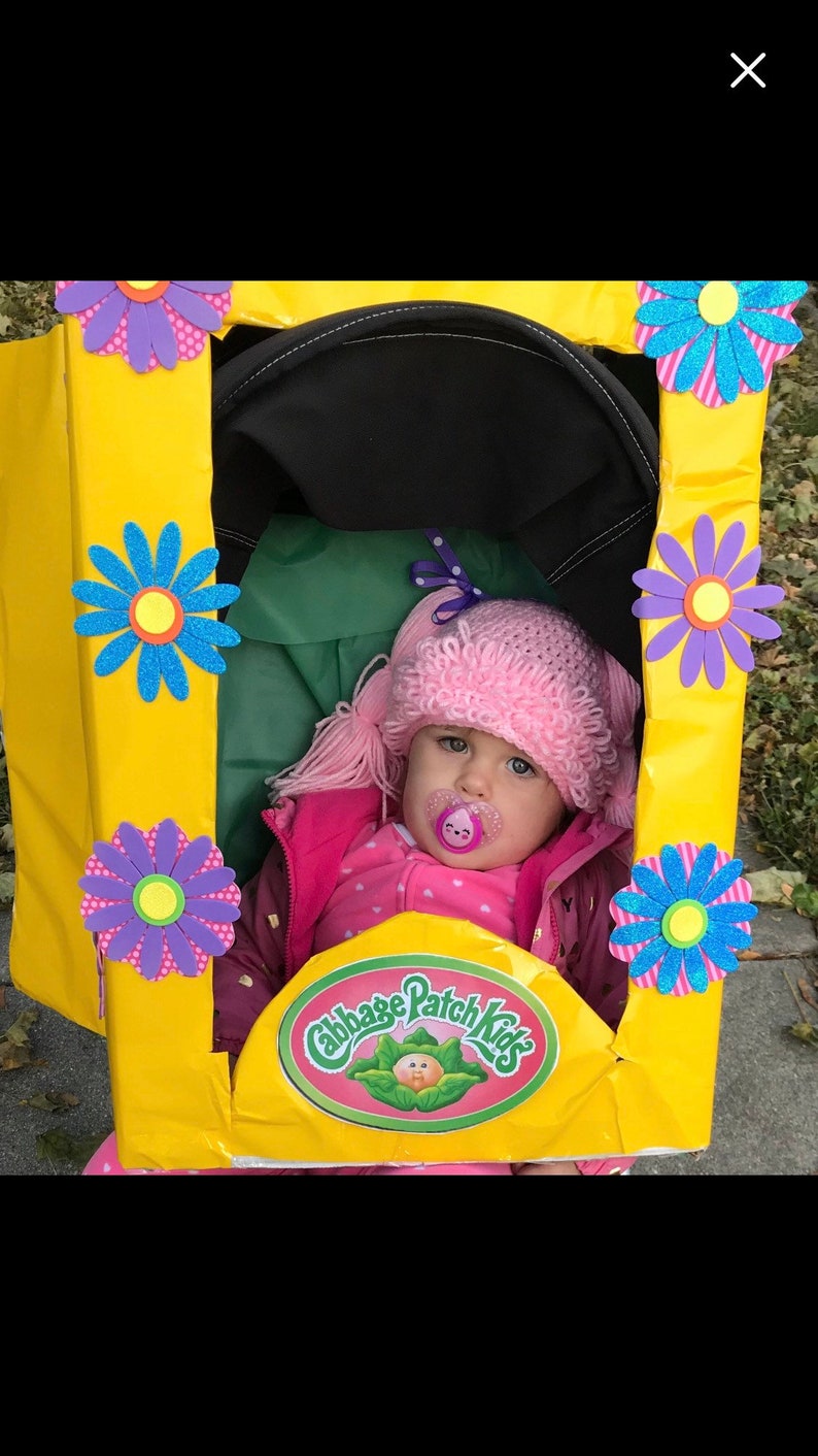K&ouml;nnte beinhalten: Ein Baby, das als Cabbage Patch Kid Puppe verkleidet ist. Das Baby tr&auml;gt eine rosa H&auml;kelm&uuml;tze mit Z&ouml;pfen, ein rosa-wei&szlig;es Hemd und eine rosa Jacke. Das Baby sitzt in einem gelben Pappkarton, der mit Blumen und dem Cabbage Patch Kids Logo verziert ist.