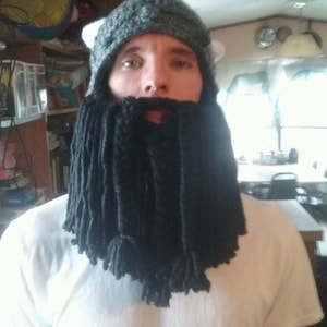 May include: A person wearing a gray and white knitted hat with a long black yarn beard.