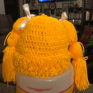 K&ouml;nnte beinhalten: Gelber H&auml;kelhut mit zwei langen Z&ouml;pfen und wei&szlig;en Schleifen. Der Hut hat einen flauschigen Saum am unteren Rand.