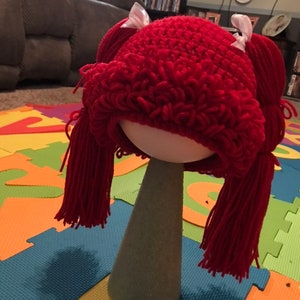 K&ouml;nnte beinhalten: Eine rote geh&auml;kelte Puppenper&uuml;cke mit zwei Z&ouml;pfen und wei&szlig;en Schleifen. Die Per&uuml;cke sitzt auf einem Styropor-Kopf.