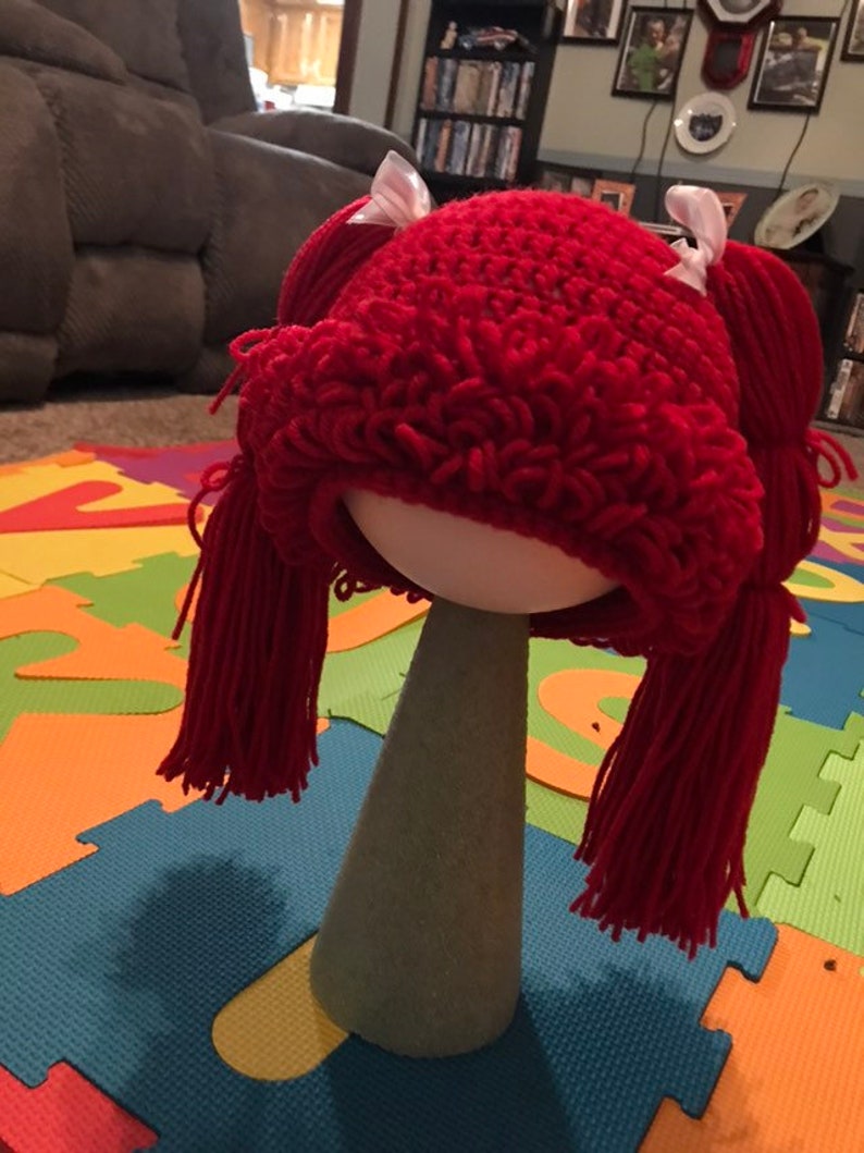 K&ouml;nnte beinhalten: Eine rote geh&auml;kelte Puppenper&uuml;cke mit zwei Z&ouml;pfen und wei&szlig;en Schleifen. Die Per&uuml;cke sitzt auf einem Styropor-Kopf.