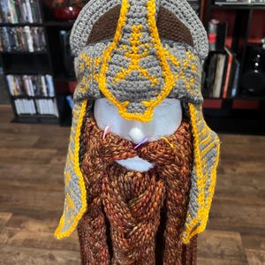 May include: A handmade crochet hat and beard set in the style of a dwarf. The hat is gray with brown and yellow accents, and the beard is a braided brown and orange. The set is designed to be worn as a costume.