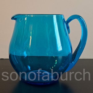 May include: A bright blue glass pitcher with a rounded body and a handle. The pitcher is made of clear glass with a slight blue tint.