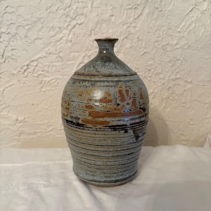 May include: A handcrafted ceramic vase with a narrow neck and rounded body. The vase features a blend of blue, brown, and black horizontal stripes and textured areas. The pottery has a rustic, artisanal appearance, suitable for home decor.