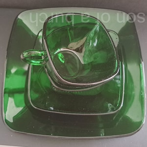 Può includere: Un set di tre piatti in vetro verde, tra cui un piatto quadrato e un piattino quadrato più piccolo con una tazza in vetro verde abbinata.