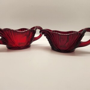 Könnte beinhalten: Zwei rubinrote Sahnekännchen aus Glas mit fächerförmigem Design. Jedes Kännchen hat einen Henkel und einen gewellten Rand. Das Glas hat eine tiefe, satte rote Farbe, und das Design verleiht den Kännchen einen Vintage-Look.