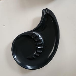 Pode incluir: Um cinzeiro preto brilhante com um design curvo único. O cinzeiro apresenta uma área circular com várias ranhuras para apoiar cigarros. Sua forma assemelha-se a um chifre ou espiral estilizada, oferecendo uma estética moderna.