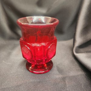 Op de afbeelding: Een kleine, rode glazen vaas met een voet en een decoratief patroon. De vaas heeft een ronde, open bovenkant en is gemaakt van helder glas met een rode tint.