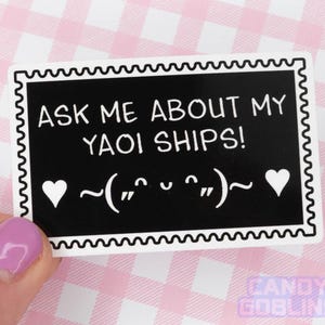 May include: A rectangular sticker with a black background and white text that reads "ASK ME ABOUT MY YAOI SHIPS!" with a heart on each side of a face symbol. The sticker has a white border with a wavy design.