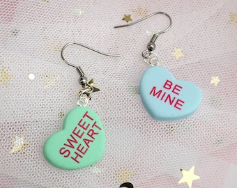 May include: A pair of heart-shaped earrings with a silver hook and a charm. One earring is light blue with the text "BE MINE" in pink. The other earring is light green with the text "SWEET HEART" in pink.