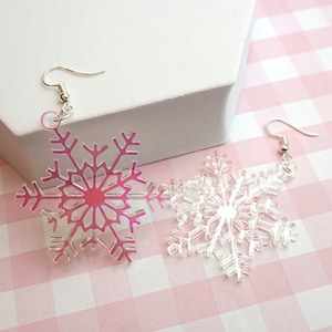 May include: A pair of snowflake earrings. One earring is a gradient of pink and clear, the other is clear. Both earrings have silver-coloured fish hook ear wires. The earrings are on a pink and white checkered surface.