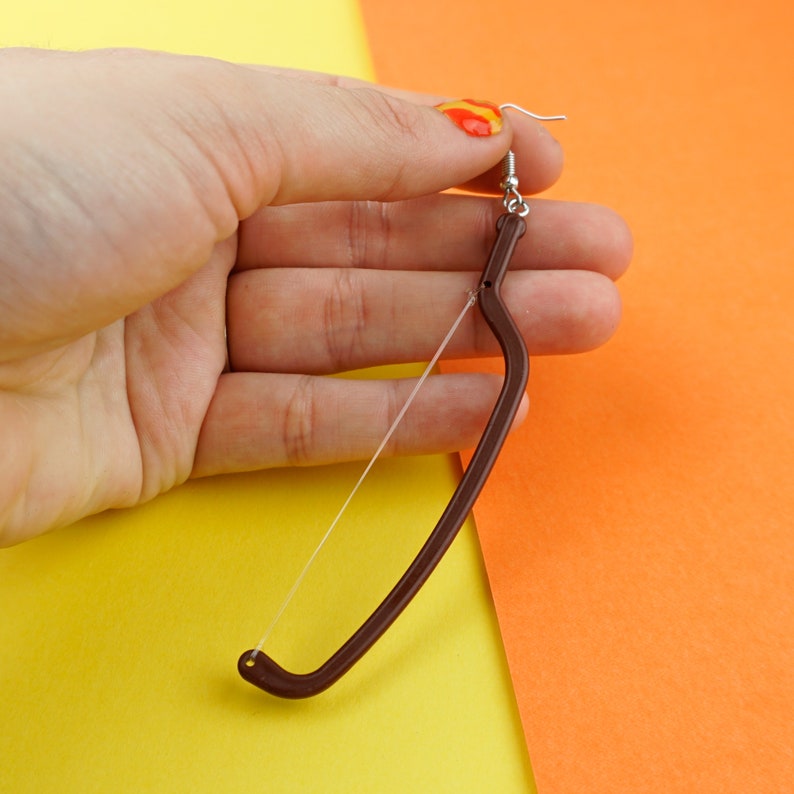 May include: A brown bow and arrow earring with a silver hook. The earring is held in a hand with a red nail polish.