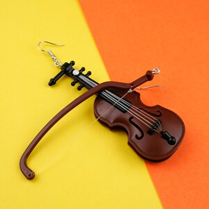 May include: A brown violin-shaped earring with a black bow and silver hook. The earring is on a yellow and orange background.