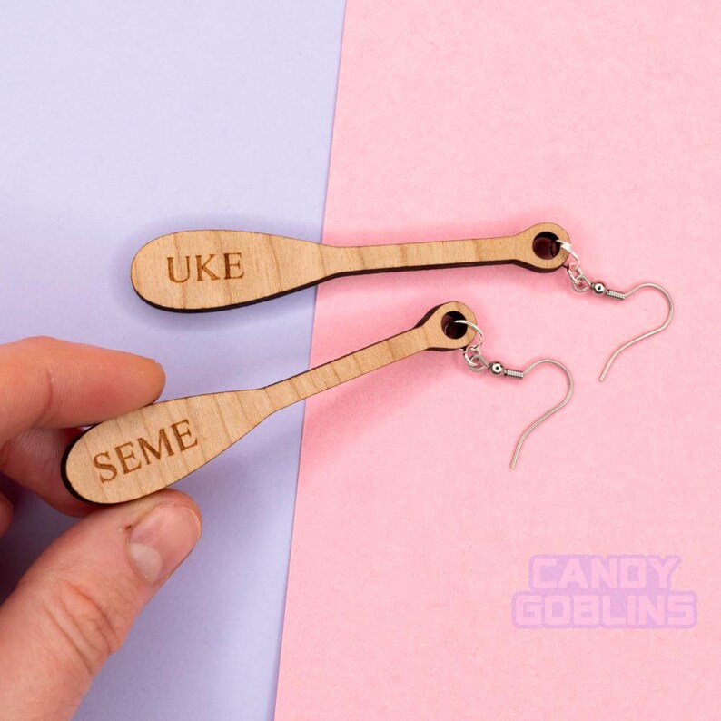 May include: A pair of wooden earrings shaped like paddles. One earring is engraved with the word "UKE" and the other is engraved with the word "SEME".