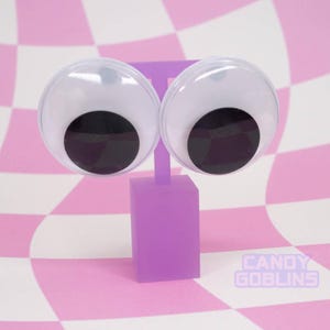 以下が含まれることがあります： 白い虹彩と黒い瞳を持つ、大きなギョロ目イヤリングのペア。紫色のスタンドに取り付けられています。背景はピンクと白のチェック柄です。「CANDY GOBLINS」の文字が右下に表示されています。ユニークなアクセサリー。