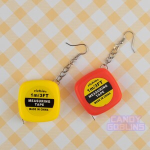 Puede incluir: Un par de pendientes con cintas métricas en miniatura de color amarillo y naranja con el texto "richies 1m/3FT MEASURING TAPE MADE IN CHINA" en cada una.