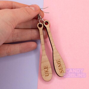 May include: A pair of wooden earrings shaped like paddles. The earrings are engraved with the words "UKE" and "SEME".