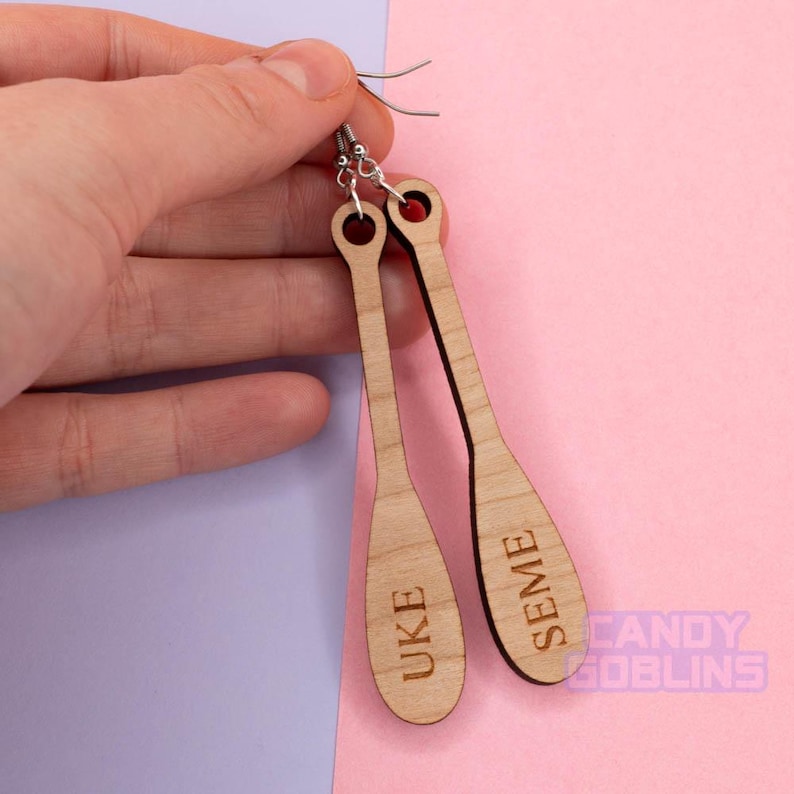 May include: A pair of wooden earrings shaped like paddles. The earrings are engraved with the words "UKE" and "SEME".