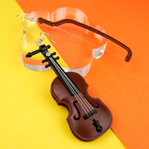 May include: A pair of earrings featuring a brown violin-shaped charm with black details and a silver hook. The earrings are displayed on a yellow and orange background.