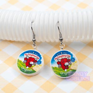A pair of Laughing Cow earrings lie against a white photo prop