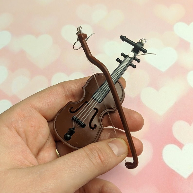 May include: A pair of earrings shaped like a violin and bow. The violin is brown with black strings and the bow is brown with a silver hook.