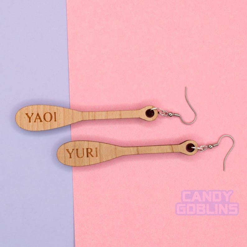 May include: A pair of wooden earrings with the words "YAOI" and "YURI" engraved on them. The earrings are a light brown color and have silver hooks.