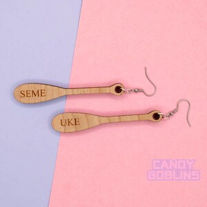May include: A pair of wooden earrings shaped like paddles. The top earring has the word "SEME" engraved on it, and the bottom earring has the word "UKE" engraved on it.