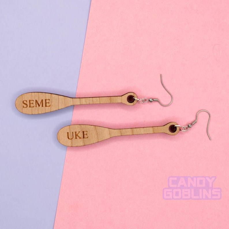 May include: A pair of wooden earrings shaped like paddles. The top earring has the word "SEME" engraved on it, and the bottom earring has the word "UKE" engraved on it.