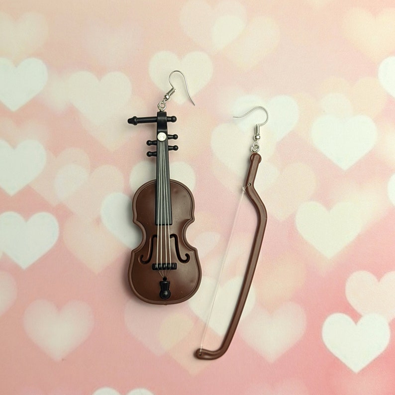 May include: A pair of earrings featuring a brown violin and a silver bow. The violin is detailed with black strings and a silver tuning peg.