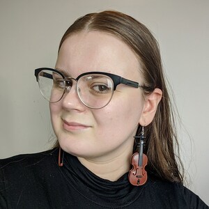 May include: A woman wearing black framed glasses and a black shirt with a pair of brown violin earrings.