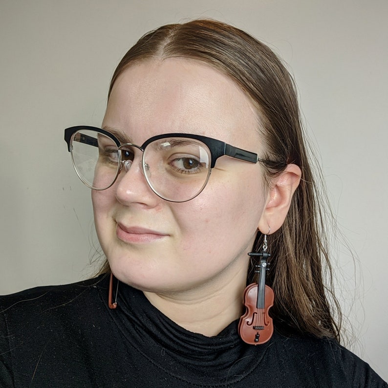 May include: A woman wearing black framed glasses and a black shirt with a pair of brown violin earrings.
