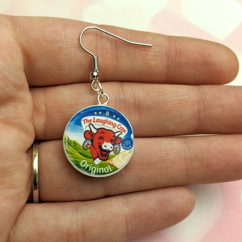 A hand holds a single Laughing Cow earring, showing the scale