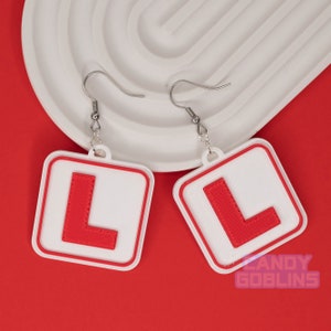 May include: A pair of white earrings with red L-shaped charms. The charms are shaped like learner driver signs.
