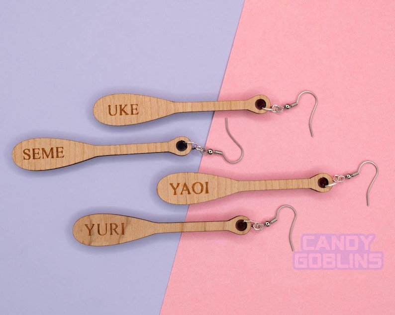May include: Four pairs of wooden earrings with silver hooks. The earrings are shaped like paddles and have the words "Uke", "Seme", "Yaoi", and "Yuri" engraved on them.