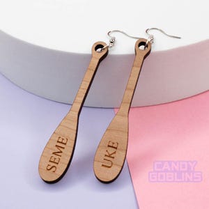 May include: A pair of wooden earrings shaped like paddles. The left earring is engraved with the word "SEME" and the right earring is engraved with the word "UKE".