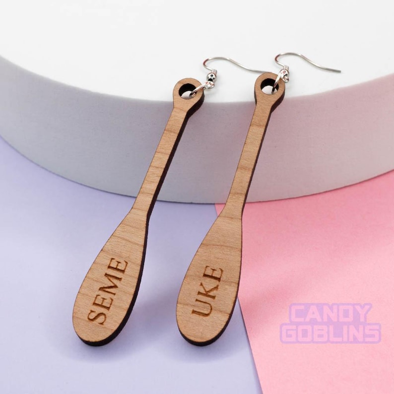 May include: A pair of wooden earrings shaped like paddles. The left earring is engraved with the word "SEME" and the right earring is engraved with the word "UKE".