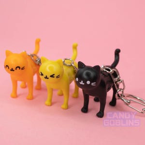 May include: Three cat-shaped keychains in orange, yellow, and black. Each cat has a unique facial expression and is attached to a silver chain. The keychains are standing on a pink surface. The text "CANDY GOBLINS" is visible.